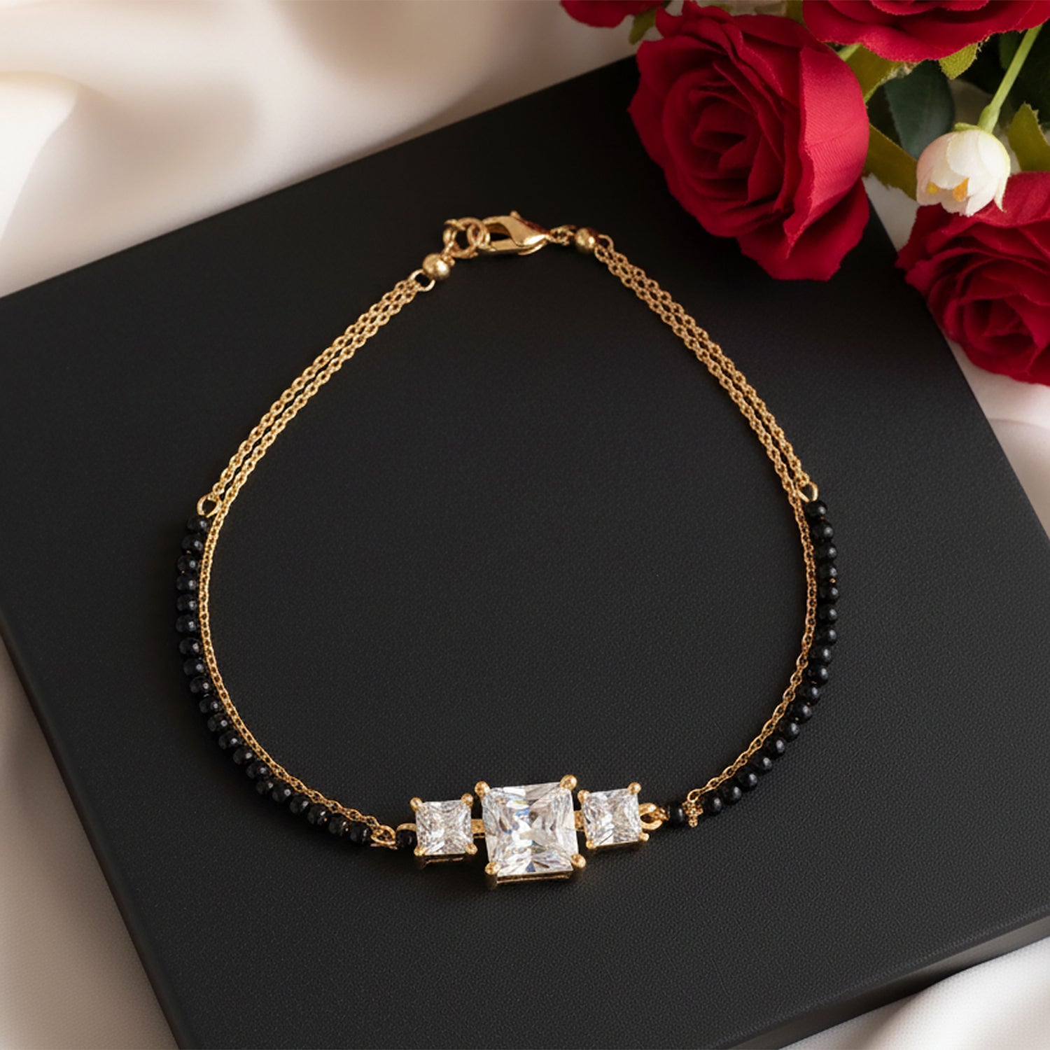 Elegant black beads mangalsutra gold bracelet featuring three square sparkling stones, displayed on a black box with red roses in the background.