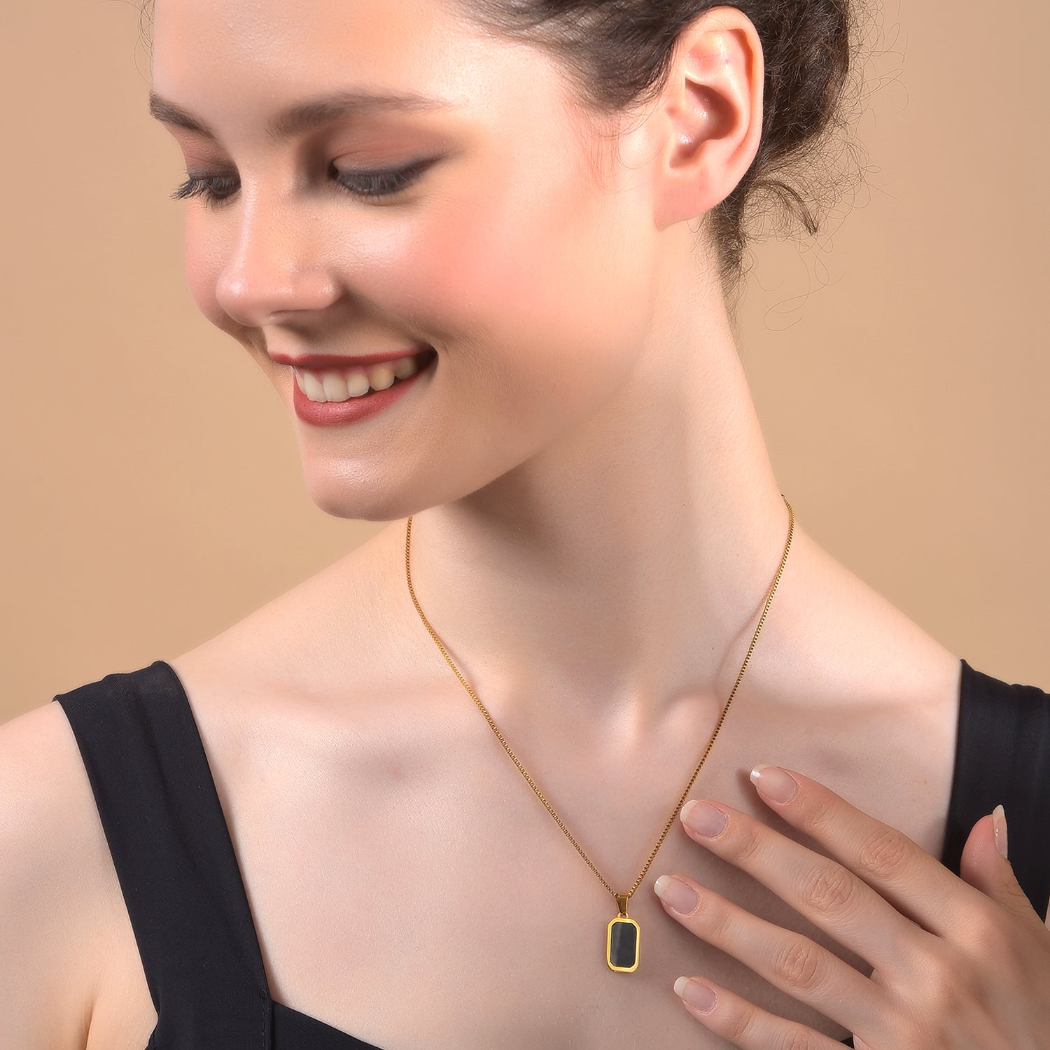 Close-up of a person wearing a minimalist gold necklace design featuring a thin gold box chain and a small rectangular black onyx pendant.