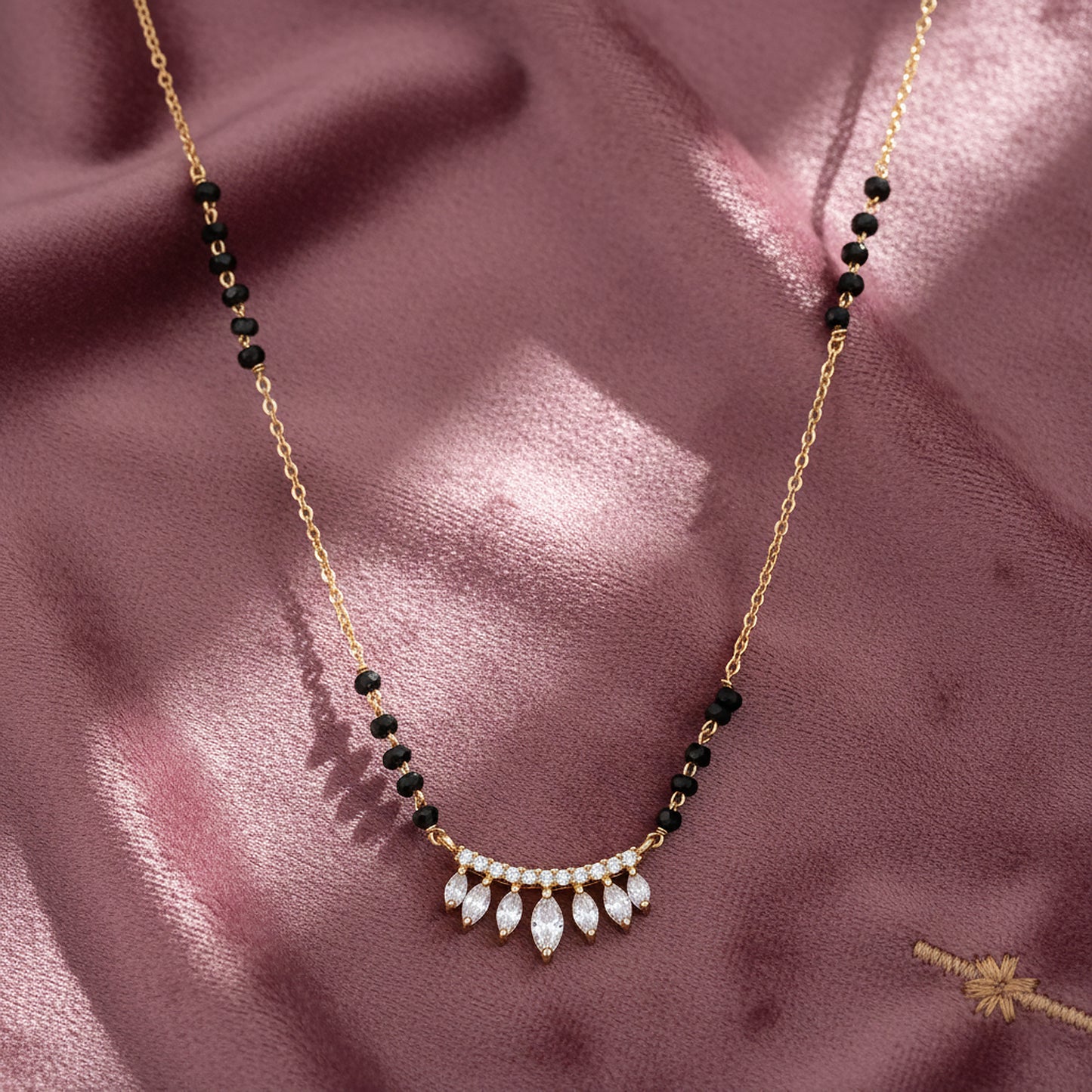 Gold mangalsutra with black beads and a curved pendant featuring sparkling leaf-shaped CZ stones, displayed on a soft satin fabric background