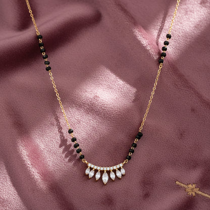 Gold mangalsutra with black beads and a curved pendant featuring sparkling leaf-shaped CZ stones, displayed on a soft satin fabric background