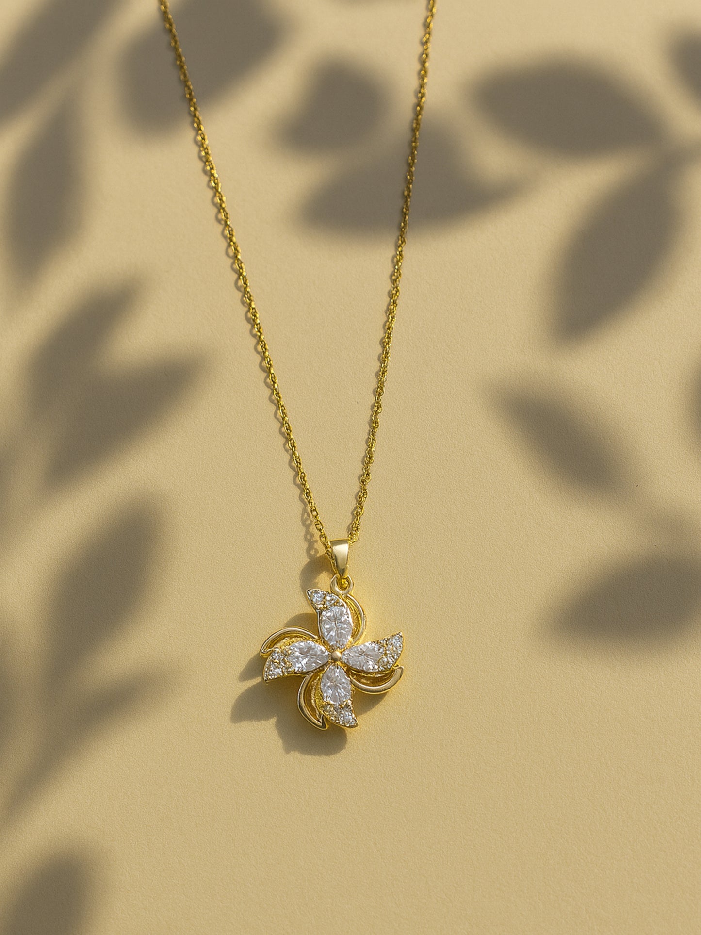 Gold necklace for women with a shimmering four-petal floral pendant, shown on a neutral tan background with soft leaf shadows.