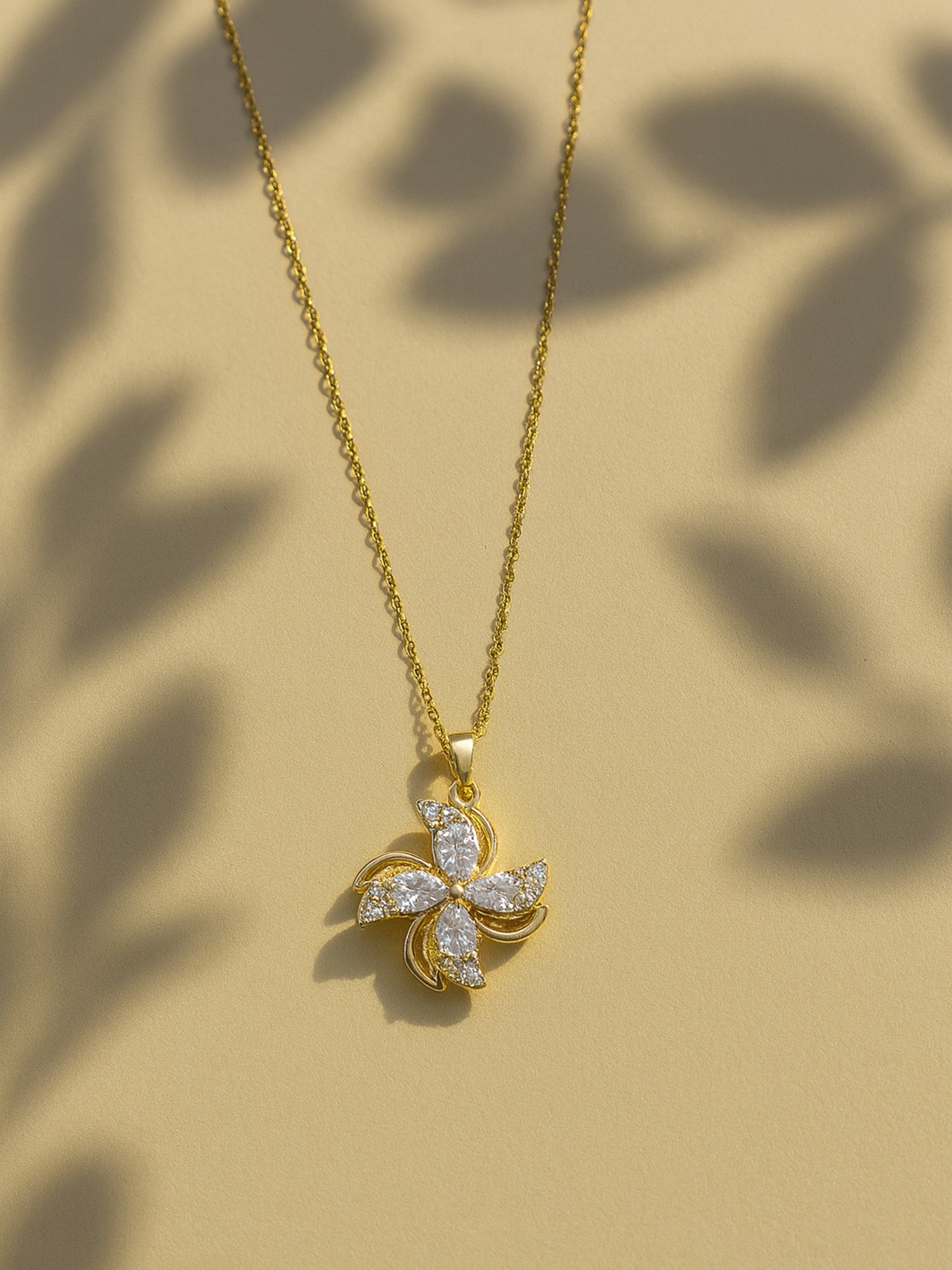 Gold necklace for women with a shimmering four-petal floral pendant, shown on a neutral tan background with soft leaf shadows.