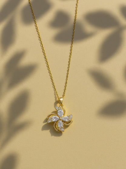 Gold necklace for women with a shimmering four-petal floral pendant, shown on a neutral tan background with soft leaf shadows.