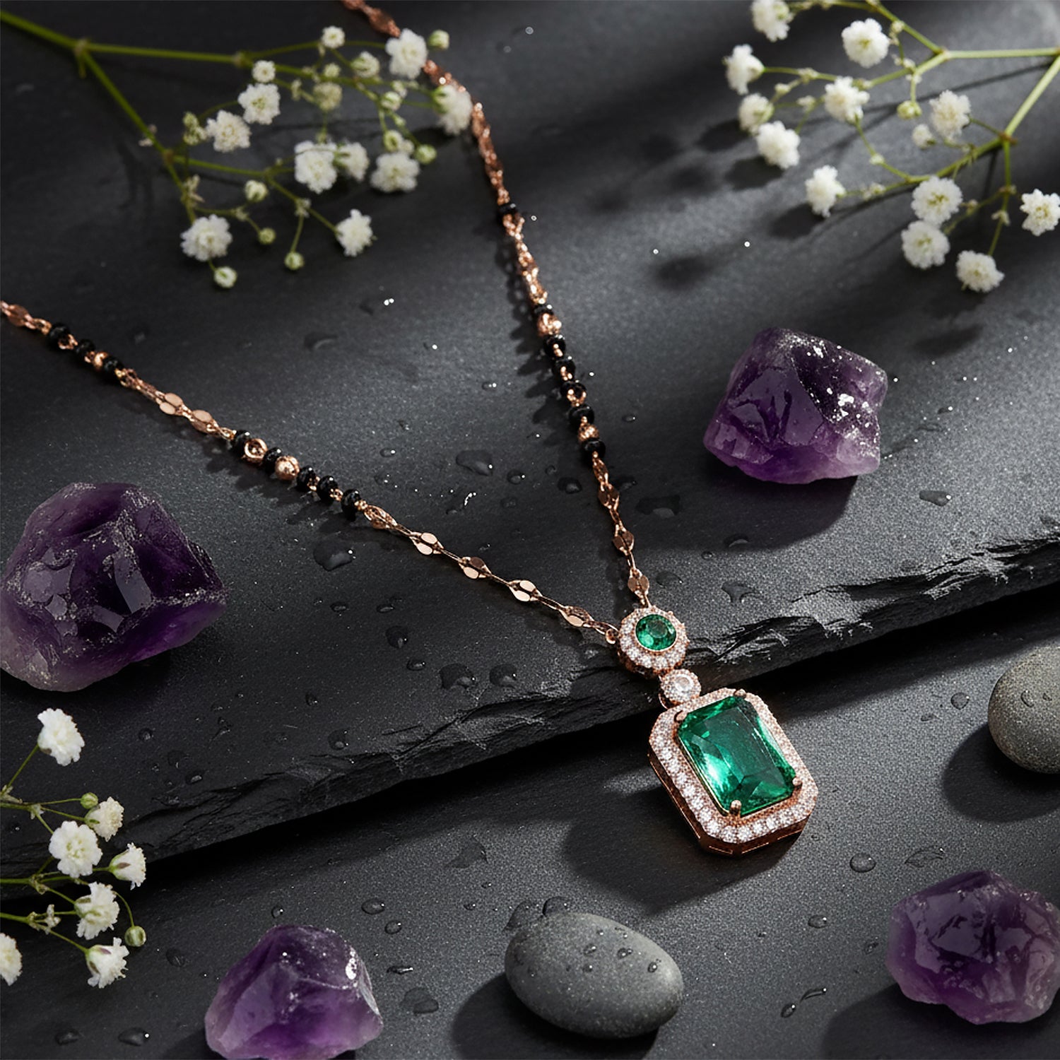 Fancy short mangalsutra design with an emerald pendant displayed on wet black stone with purple crystals and white flowers