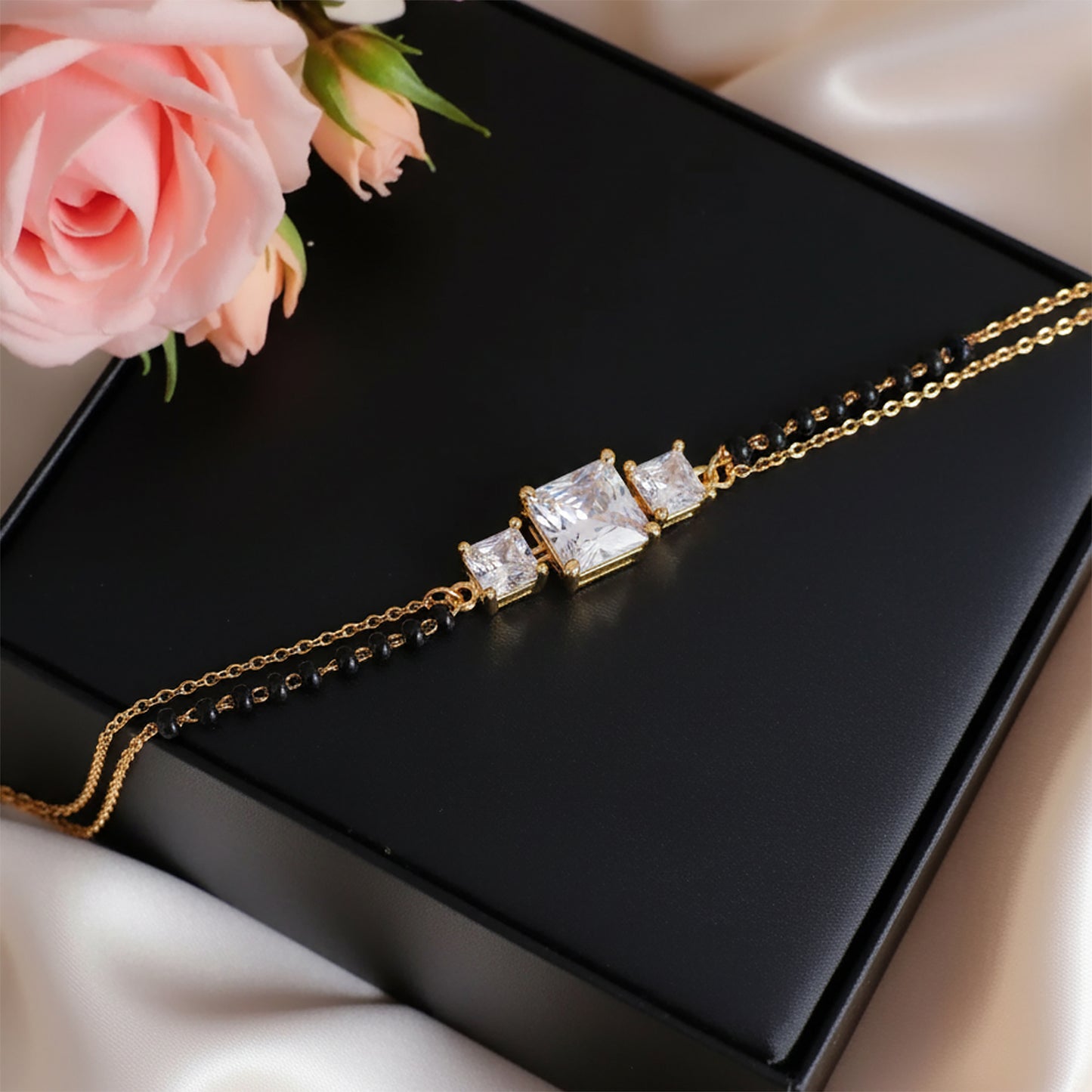 Elegant mangalsutra bracelet design featuring a gold chain with black beads and three sparkling square stones, displayed on a black box with soft pink roses.