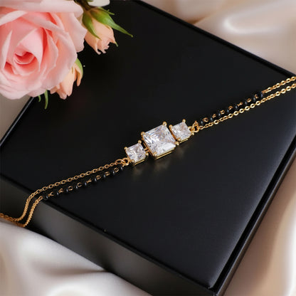 Elegant mangalsutra bracelet design featuring a gold chain with black beads and three sparkling square stones, displayed on a black box with soft pink roses.