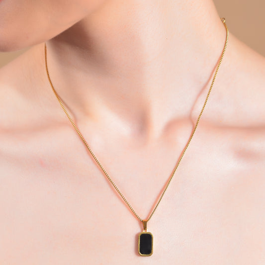 Close-up of a person wearing a minimalist gold necklace design featuring a thin gold box chain and a small rectangular black onyx pendant.