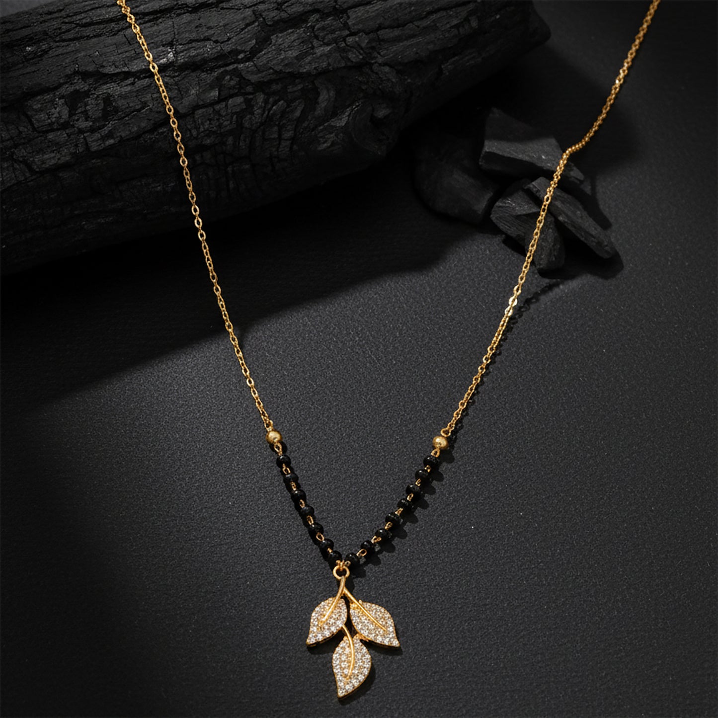 Gold plated mangalsutra design with black beads and a crystal leaf pendant on a dark textured background