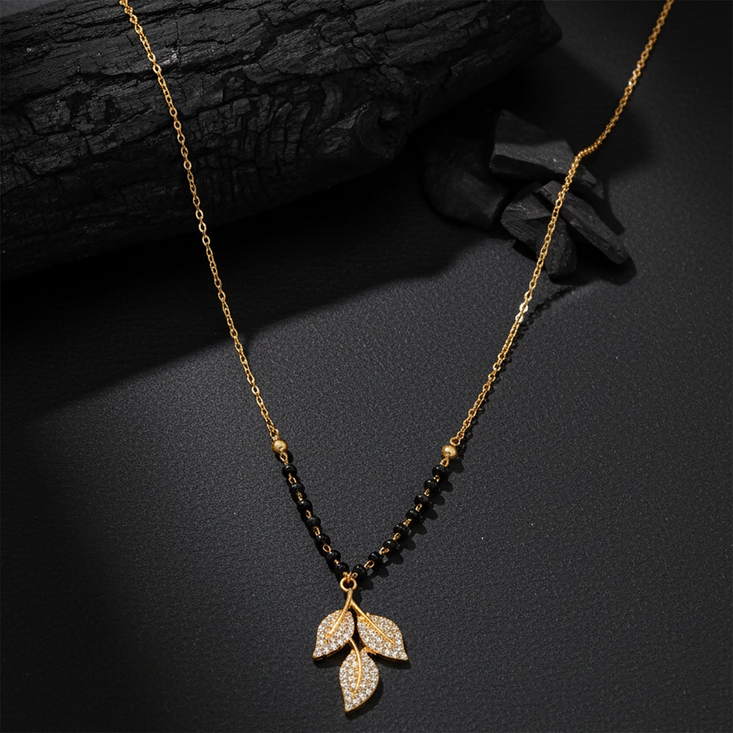 Gold plated mangalsutra design with black beads and a crystal leaf pendant on a dark textured background